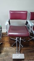 Front view of a vintage hairdressing chair with burgundy vinyl seat and backrest, metal armrests and chrome base.