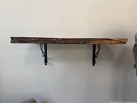 Side view of unfinished live edge wood slab showing natural bark edge and thickness, mounted on two black metal wall brackets attached to wall.