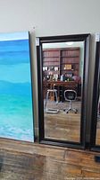 Front view of the full length wall mounted mirror leaning against a wall, next to a large painting and showing reflections of chairs and bookshelves.