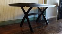 Side view of the black wooden multitask table showing X-shaped legs and some scratches on edge of tabletop.