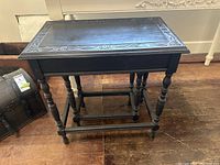 Front view showing larger nesting table with carved wood trim and turned legs, placed on wooden floor next to wooden chest