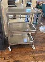 Full front view of stainless steel three-shelf rolling cart showing horizontal bars on one side and wheels.