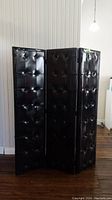 Three tall, black vinyl padded room divider panels shown folded and standing upright in a room with wood flooring and white wall paneling.