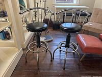 Pair of two chrome metal swivel bar stools with circular black vinyl seats and curved chrome backs, sitting on a wooden floor inside a room with other furniture.