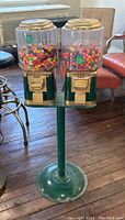 Front and side view of vintage dual canister gum-ball candy machine filled with colorful candy, mounted on green metal stand with coin mechanisms and missing finials on canister tops.
