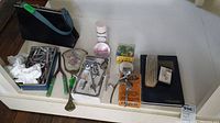 Photo of the vintage grooming items arranged on a white shelf showing razors, hair curlers, handheld mirror, purse, powder container, colorful beads, wooden hairbrush, book and more.