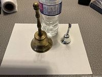 Two hand bells side by side with a water bottle for scale. One bronze, one silver-tone.