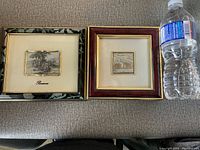 Two framed silver miniature pictures with a water bottle for scale