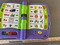 Open purple plastic interactive ABC musical toy book showing page with letters U to Y on left page, letters Z to O on right page with corresponding words and colorful illustrations.