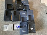 Top-down view of multiple battery chargers including Canon, Sony, Panasonic Lumix and unbranded chargers with several camera batteries laid out in front.