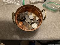 Top-down view of copper jar filled with a variety of coins of different sizes and metals, placed on a fabric surface with additional bags visible in the surrounding area.