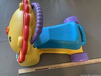 Side view of Fisher Price lion-shaped ride-on toy with blue seat and purple wheels, and visible purple handlebar.