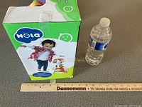 Front of box showing toddler riding the toy and brand logo, next to a water bottle and ruler for size comparison.
