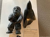 Two dark green-black soapstone carvings (standing human figure and bird) with certificate tags on paper background