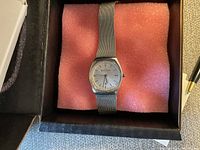 Photo of a silver-toned Skagen women's wristwatch with a mesh metal band. The watch face is missing the glass cover revealing the dial directly. Watch is placed on pink foam inside a black box.