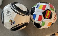Two deflated soccer balls side by side on carpeted floor: Adidas Jabulani 2010 World Cup edition and another with international flags.