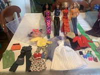 Four vintage Barbie dolls lined up wearing various outfits with accessories laid out around them on a table.