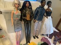 Three Barbie dolls and one Ken doll standing in front of a storage container, dressed in various fashion wear.