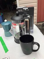 Bodum French press coffee maker with original box, Egan stove top espresso maker, and black ceramic mug arranged on table.