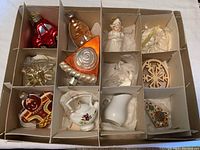 Twelve Christmas ornaments made from glass, ceramic, and wood, neatly arranged in a segmented storage box. Ornaments include a red vehicle, bird, shell, angels, star, gingerbread man, teapot, pitcher, and wooden designs.
