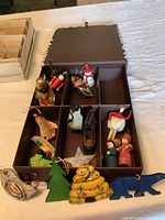 Photo showing twelve assorted Christmas ornaments including a green felt tree, yellow beehive, blue bear, animals, and figures arranged in a brown compartmentalized box.