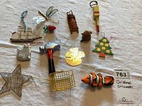 Photo showing 13 assorted Christmas ornaments including a clownfish, hammer, Christmas tree, star, dragonfly, sailboat, cat, basket, strawberry, and other small wooden and metal ornaments on a white cloth background.