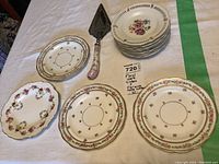 Seven Dresden cake plates with pierced edges and gold trim stacked with front rose patterned plates and cake cutter arranged around
