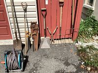 All garden tools arranged standing upright including saws, shovel, spade, fork, shears and hose reel.