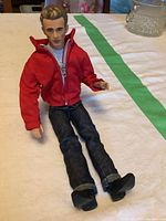 Full view of the Mattel James Dean doll seated on a white surface with a green stripe in the background, showing clothing and hair details.
