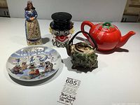 Photo shows 5 decorative collectible items including a Jim Shore grandmother figurine missing wings, an 8-inch racoon skating party plate, vintage tomato-shaped teapot, clown head condiment server, and nautilus shell teapot.