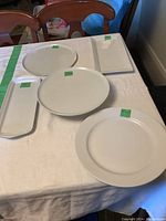 Five white porcelain serving pieces displayed on a table, including plates and trays in various shapes and sizes with visible green stickers.