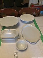 Photo showing all five porcelain serving pieces arranged on a table including a lidded rectangular dish, small bowl, round serving dish with handles, and two footed plates with scalloped edges.
