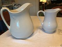 Two white vintage ironstone pitchers on a table, the larger pitcher on left shows handle with internal defect, both visible in good lighting