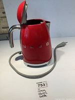 Red Smeg electric kettle with open lid showing interior, chrome handle and spout, and power cord with three-prong plug visible.