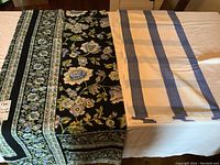 Two folded large tablecloths laid on a flat surface, one with a floral pattern on a black background and the other with blue and beige stripes on a light background.
