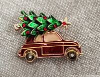 Close-up photo of a gold-tone metal and enamel brooch showing a car carrying a green iridescent holiday tree with red rhinestones and a gold star on top.