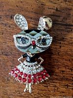 Full brooch showing entire figure of Miss Bunny posed with hands on hips, wearing rhinestone glasses and a dress decorated with red and clear rhinestones.