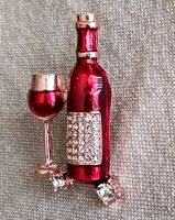 Front view of brooch showing red enamel wine bottle with rhinestone label and goblet with red enamel filled wine, on gold-tone metal base with clear rhinestone accents at base