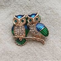 Front view of gold-tone metal owl couple brooch with colorful blue and green enamel and rhinestones on chest and eyes.