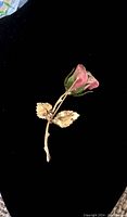 Photo of vintage pink rose brooch showing front view on black fabric background. Details of pink petals, pearl center, and gold-tone leaves and stem visible.