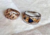 Photo showing two sterling silver rings on white fabric background. One small twisted design ring and one larger wide band ring with mixed metals and stones.