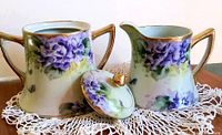 Vintage Nippon porcelain creamer and covered sugar set with hand-painted violet flowers and gold gilded details sitting on a white hand-crocheted doily.