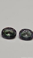 Pair of oval faceted mystic topaz gemstones showing rainbow/purple color play on white background.