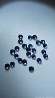 Top-down view of approximately 24 small round-cut natural blue sapphires scattered on white surface showing rich blue color and high clarity.