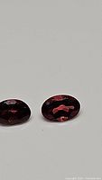 Pair of oval cut cherry red garnet gemstones on a plain surface under normal lighting showing color and facets.