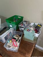 Four plastic totes filled with miscellaneous crafting materials including foam blocks, string, ribbon, and paper supplies on a wooden table against a wall