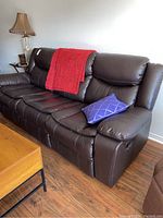 Full view of three-seat dark brown faux leather sofa with red throw blanket and blue pillow, lamp and side table in background