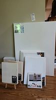 Wide photo showing multiple white stretched canvases of varying sizes stacked near a wall, some on wooden easels, including a small photo frame with a cat design in front.