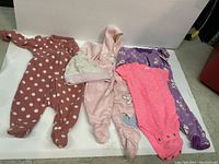 Photo showing four baby onesies and one hat laid flat on a white surface. Included are Old Navy 0-3 months polka dot zipper onesie, bebe preemie pink footed hooded onesie, purple 3-month dog print onesie, pink 3-month short sleeve onesie with hearts and buttons, and a pink and white baby hat.