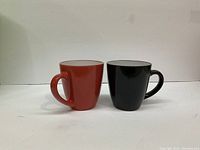Photo of two ceramic mugs, one red and one black, placed side by side on a white surface against a plain wall.
